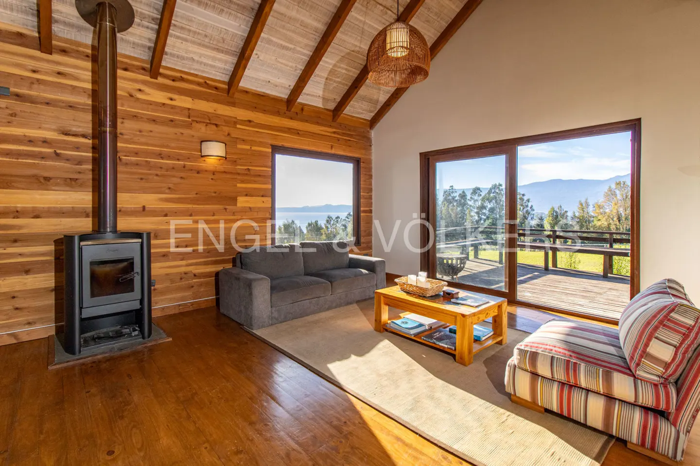 Rustic living room with wood walls, a wood-burning stove, grey sofa, striped armchair, and a view of mountains and a lake.