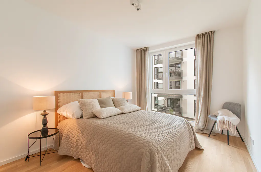 Modern bedroom with a beige quilted bed, rattan headboard, bedside lamp, and a window view of an apartment building.