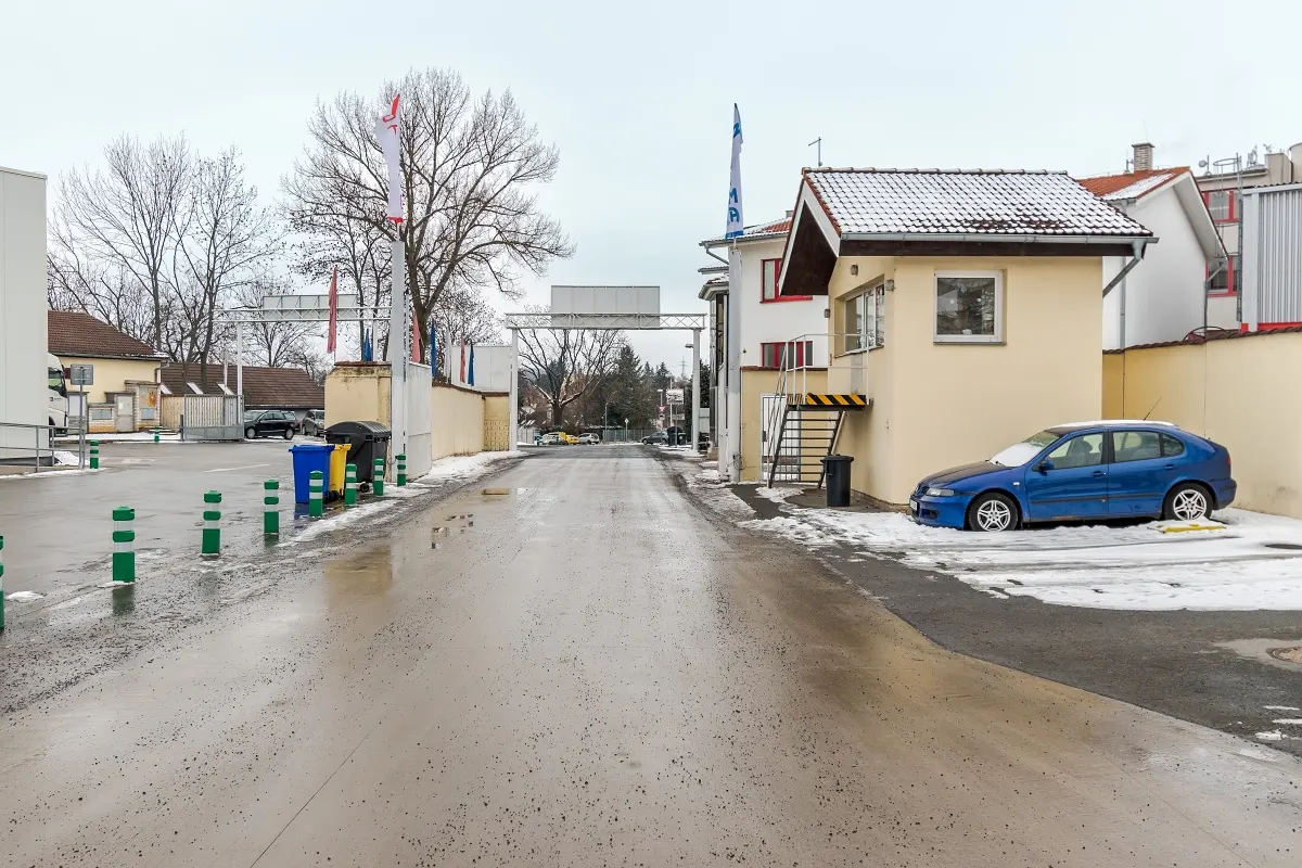 Exterior shot of a wet street with buildings, flags, and a blue car parked near a small guardhouse with a snow-covered roof.