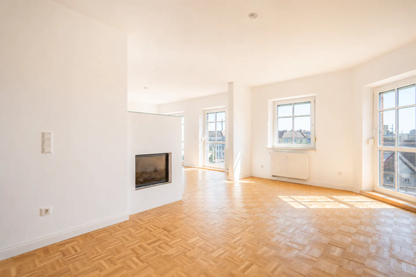 Bright, empty room with parquet floors, white walls, and a modern fireplace. Windows offer city views.