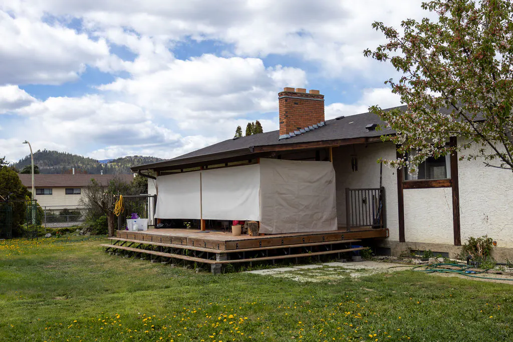 A house with a wooden deck covered by a white tarp, a brick chimney, and a green lawn with yellow dandelions.