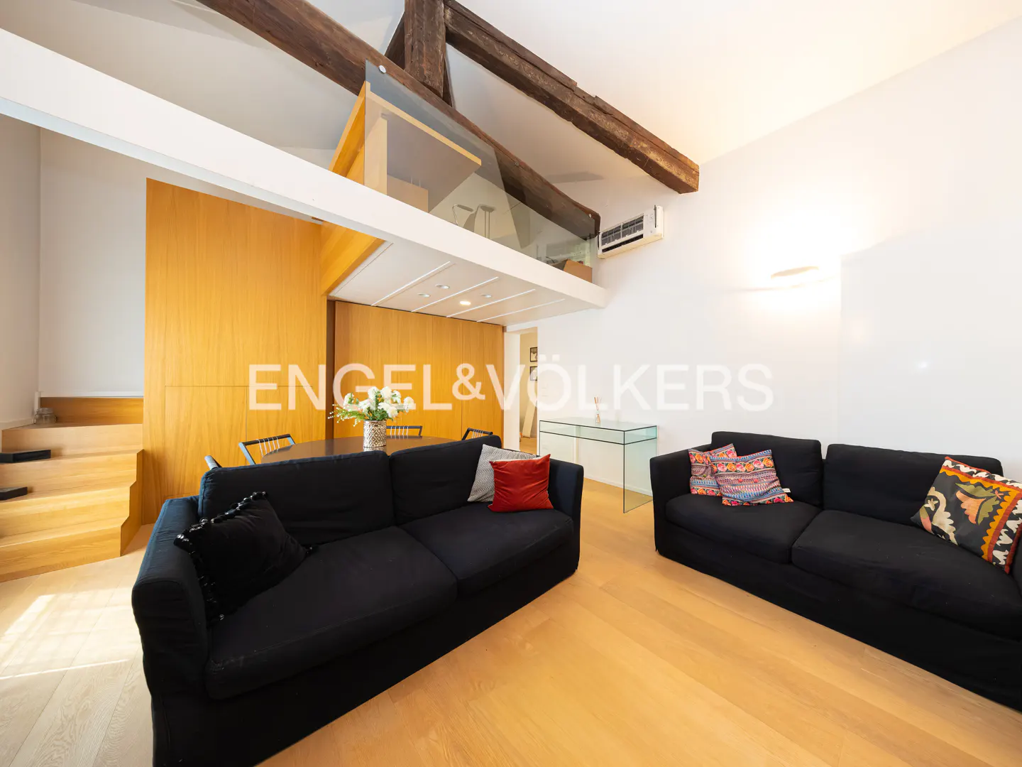 A modern living room with two black sofas, wood floors, and a glass-enclosed loft area.