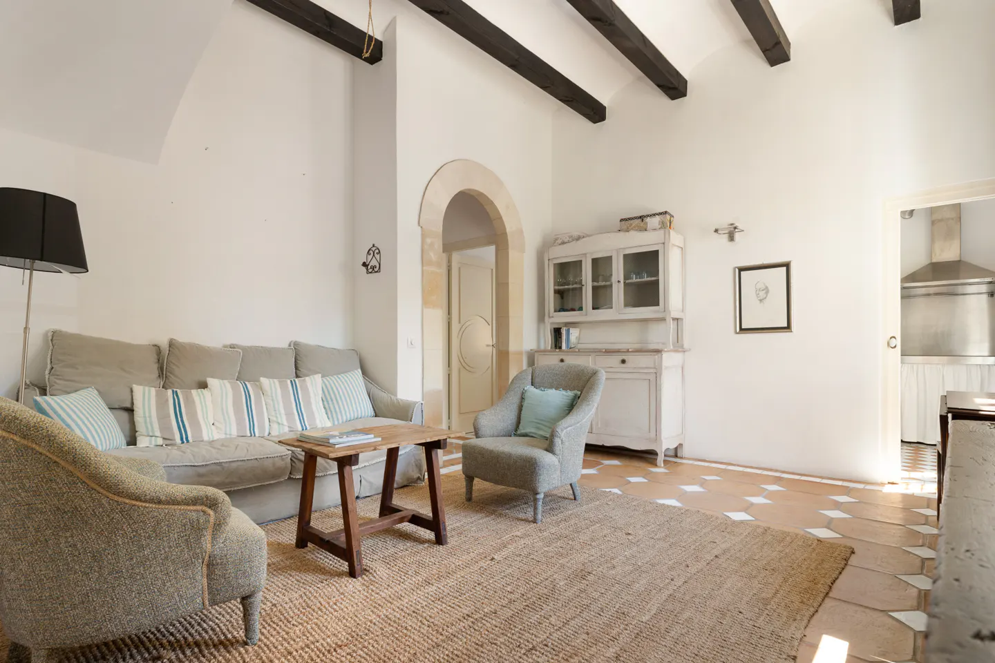 Living room with a beige sofa, two armchairs, and a wooden table on a jute rug. The walls are white, and the ceiling has dark wooden beams.