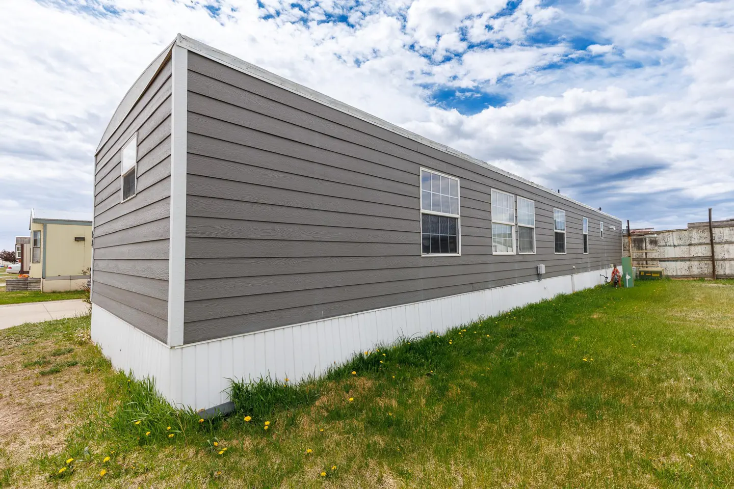 Exterior side view of a long, grey manufactured home with white skirting, set on a grassy lot under a cloudy blue sky.