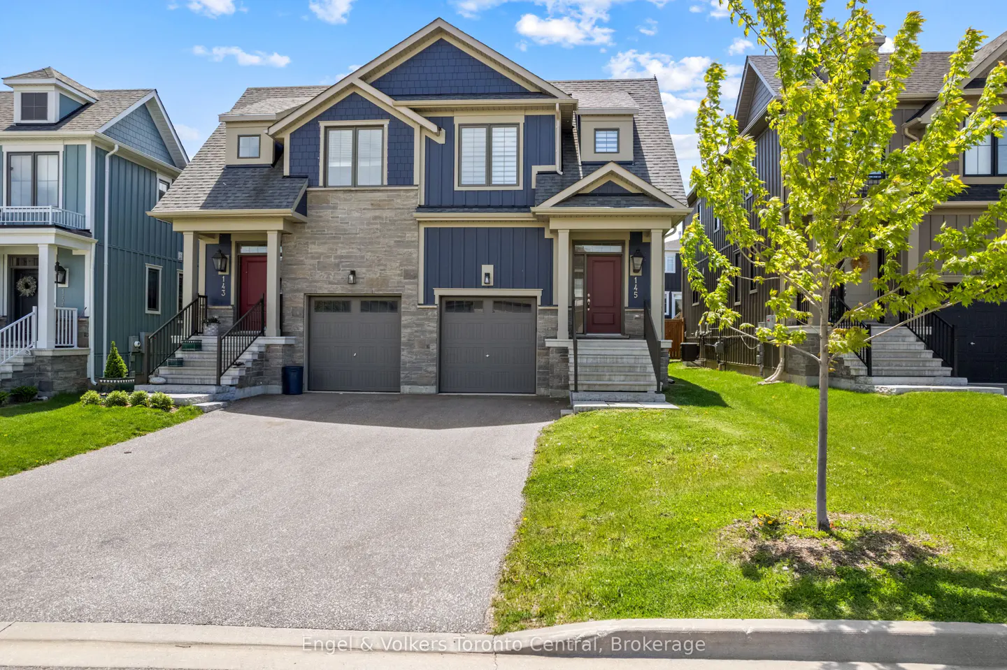 Two-story semi-detached house with blue siding, stone accents, gray garage doors, red front doors, and green lawn.