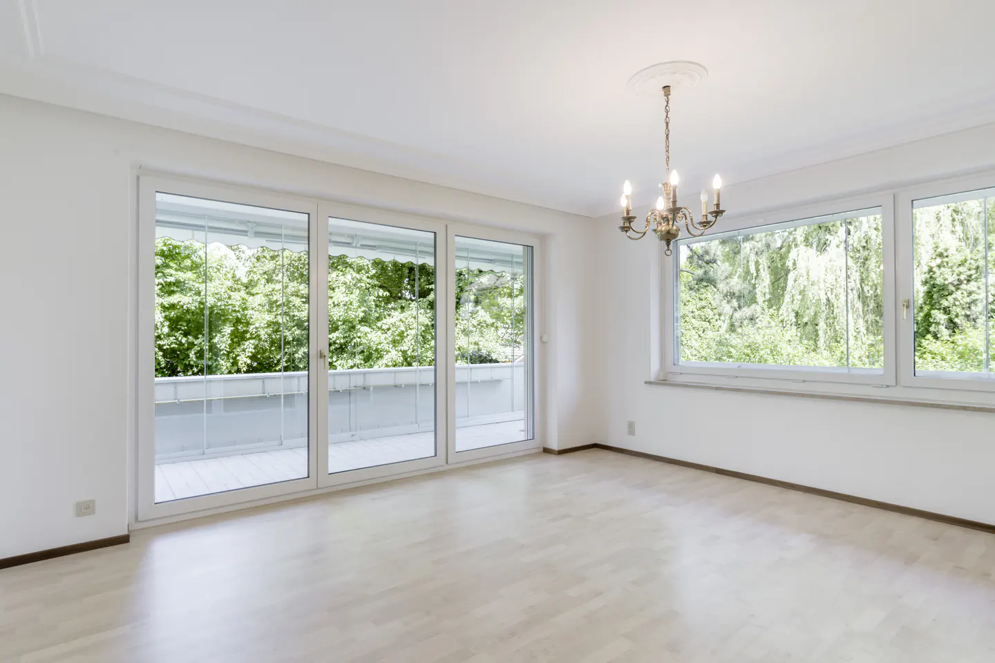Bright, empty room with light wood floors, a classic chandelier, and large windows overlooking lush green trees.