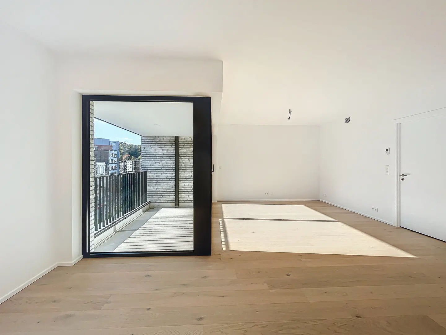 Bright, empty room with light wood floors and white walls. A black-framed sliding glass door leads to a balcony with a black railing and brick wall.