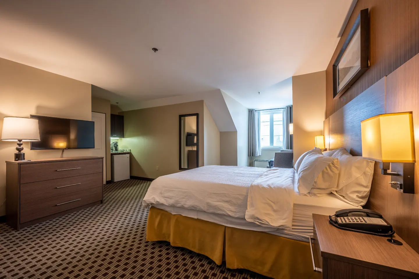 Hotel room with a queen bed, nightstand, and lamp. A TV, dresser, and kitchenette are in the background. The room has a patterned carpet.