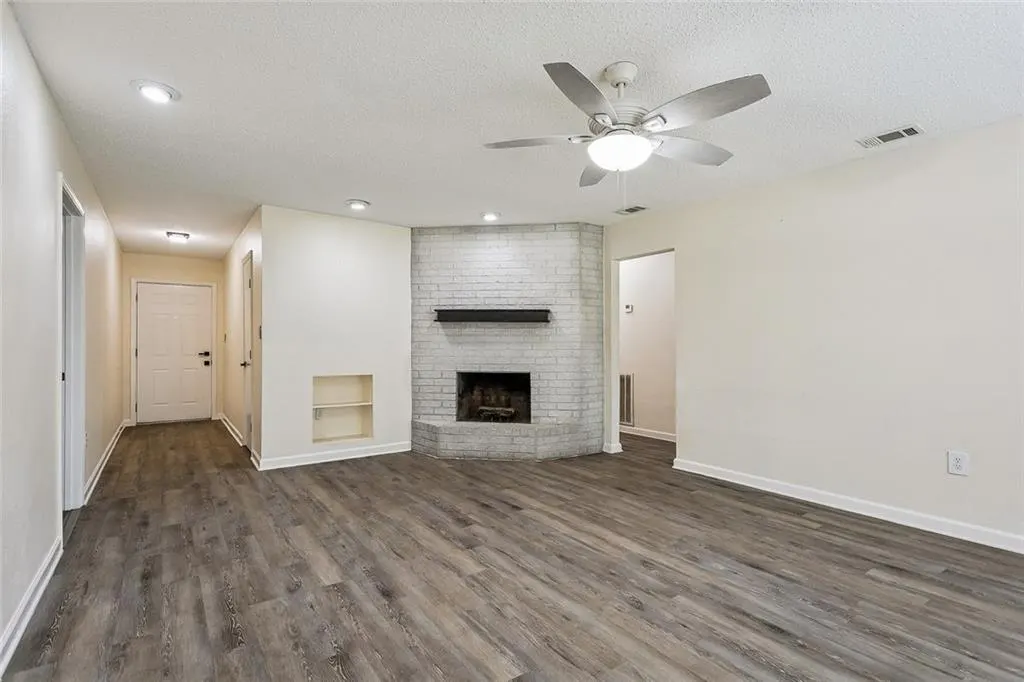 Bright living room with gray wood floors, white walls, and a brick fireplace. A ceiling fan hangs above.
