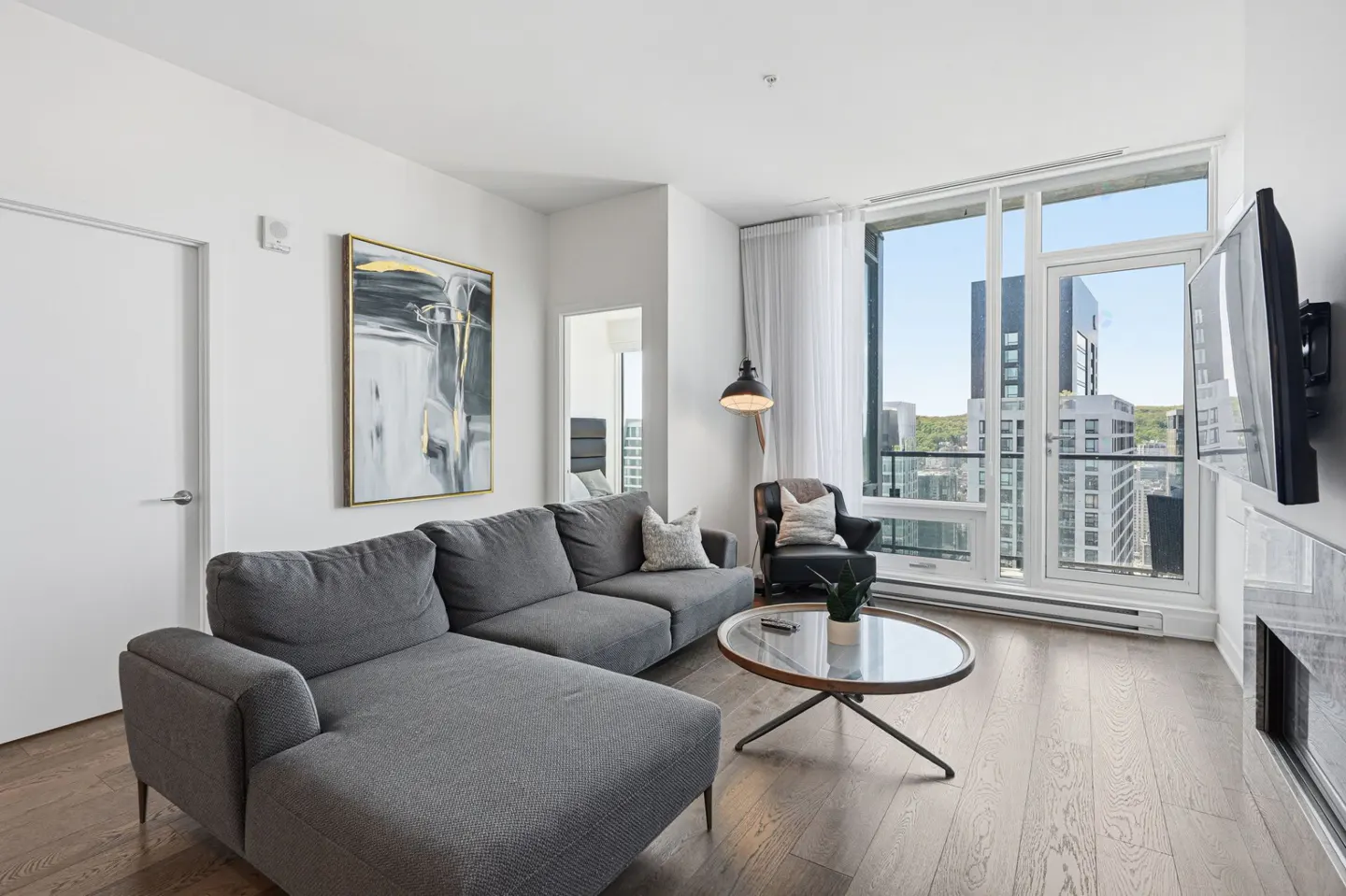 Bright living room with a gray sectional sofa, round glass table, and city view from large windows. An abstract painting hangs on the white wall.