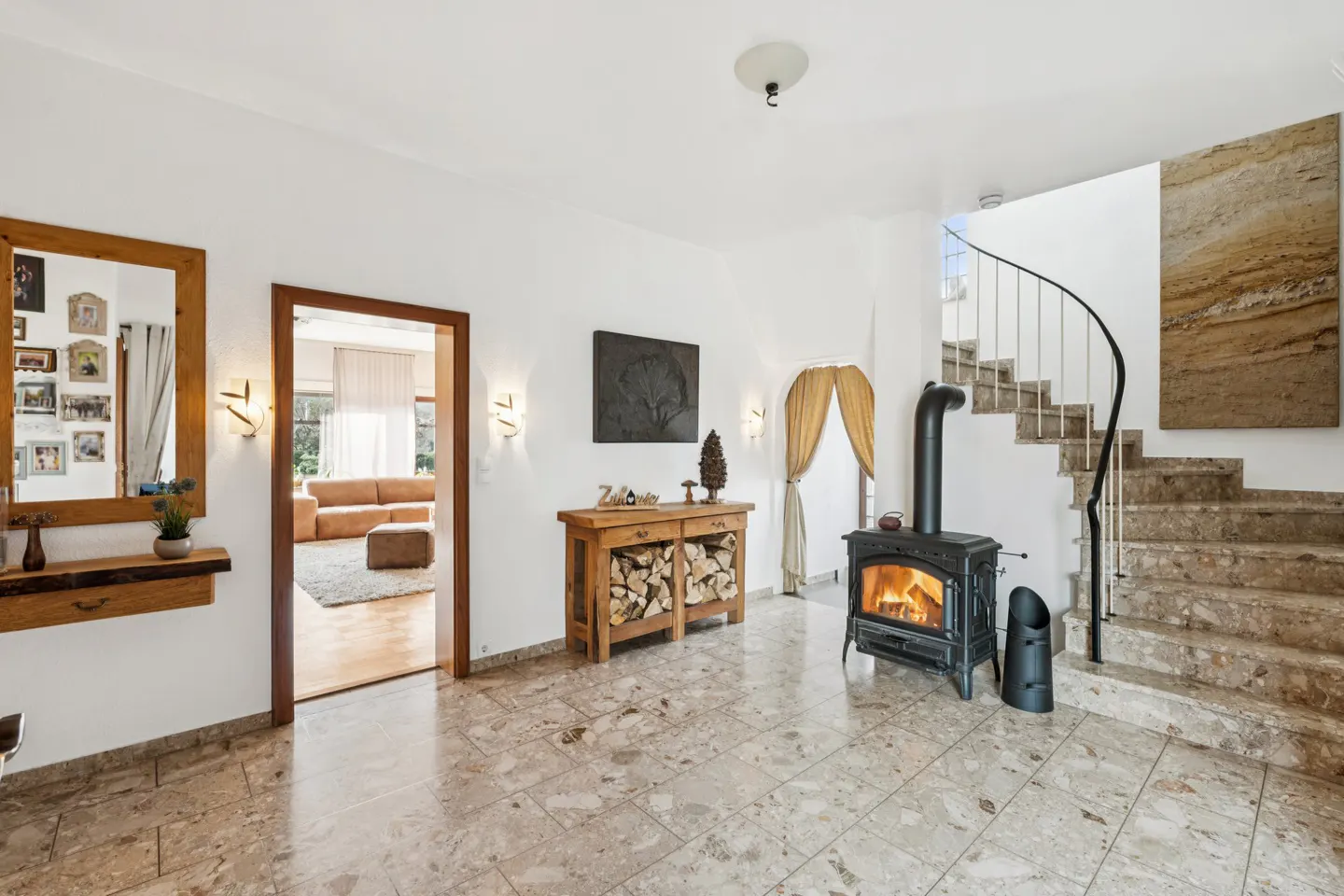 Bright foyer with marble floors, wood-burning stove, and staircase. A wood table with logs sits near the stove. A mirror and doorway are on the left.
