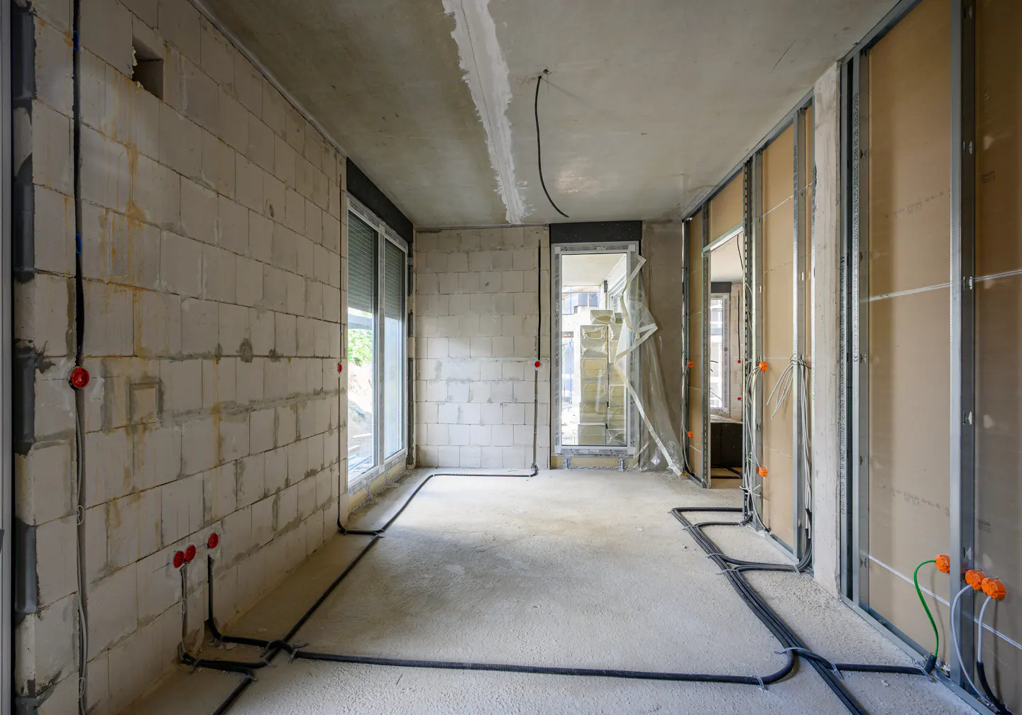 Interior view of a room under construction with concrete block walls, exposed wiring, and metal framing for drywall. Windows and a doorway are visible.