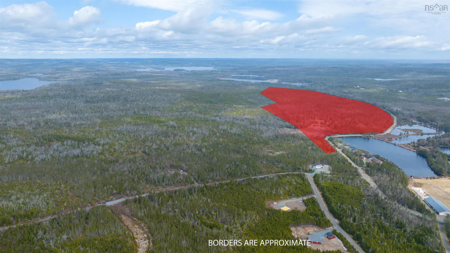 Aerial view of a wooded lot outlined in red, next to a lake and road, under a cloudy sky.
