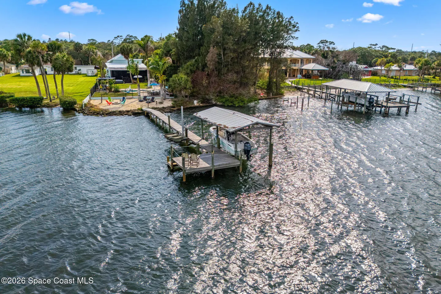 Aerial view of a waterfront property with a dock, boat lift, and a sandy beach area with colorful chairs. The water reflects sunlight.