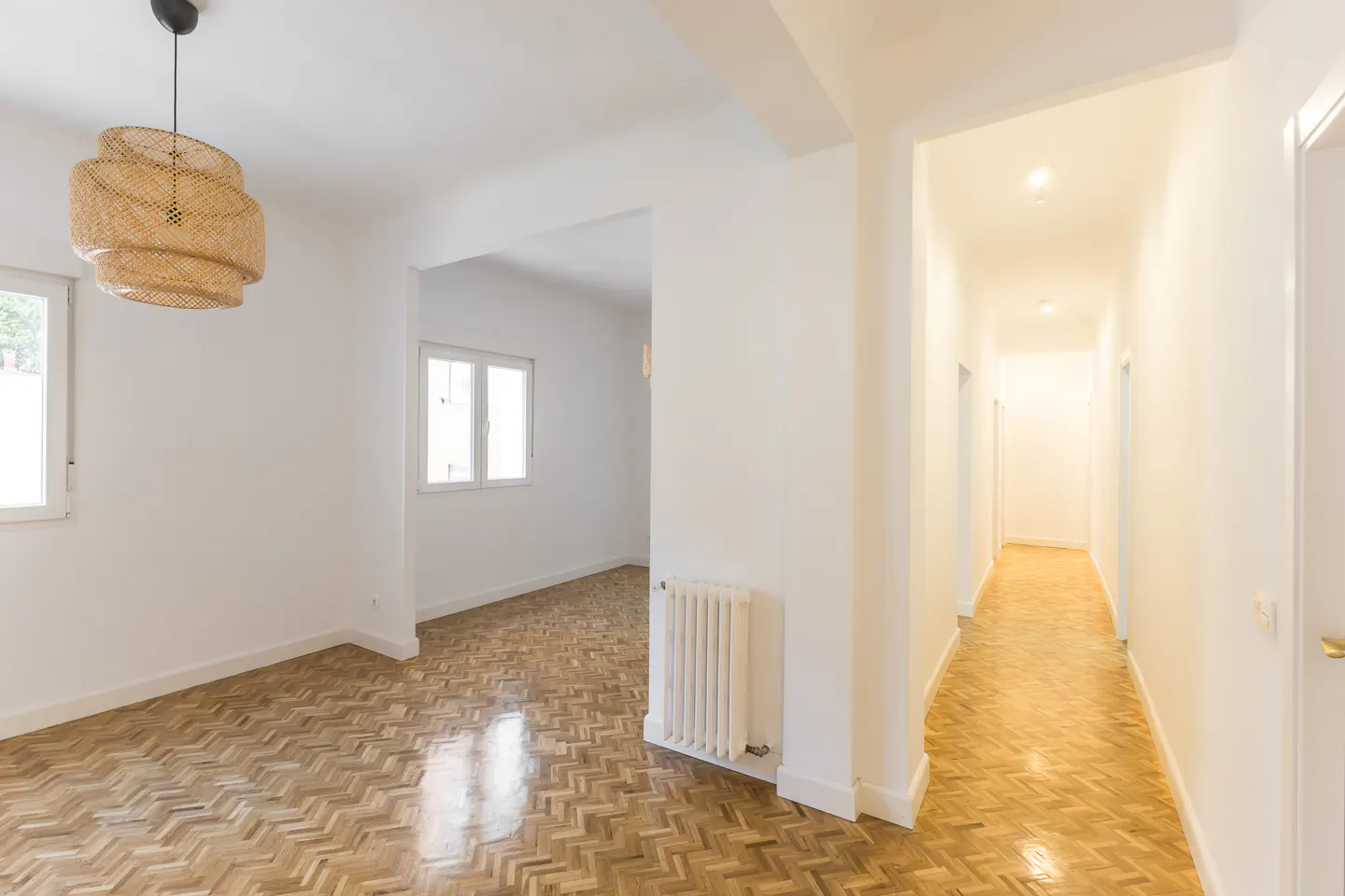 Bright, empty room with herringbone wood floors, white walls, and a woven pendant light. A hallway extends into the distance.