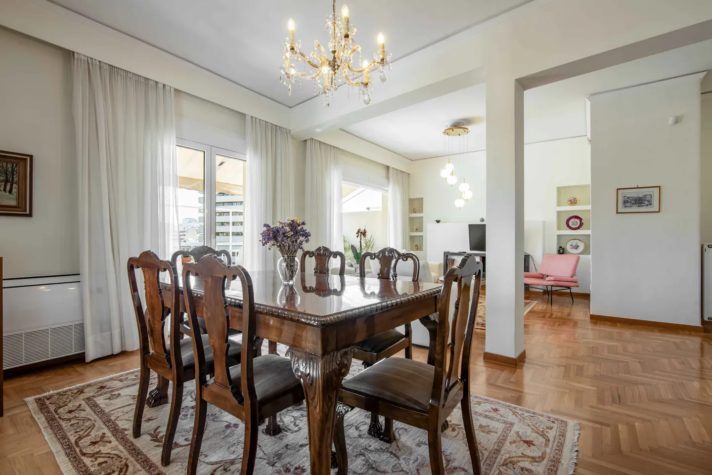 Bright dining room with a wood table, chairs, and chandelier. White curtains and a patterned rug add warmth. A glimpse of a living room is visible.
