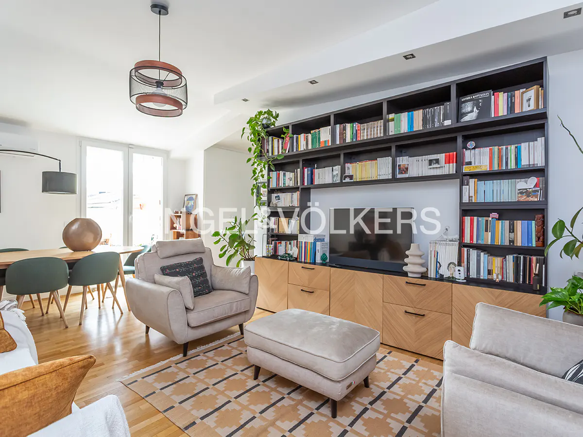 Modern living room with beige armchairs, a matching ottoman, a large wooden bookshelf with a TV, and a dining area.