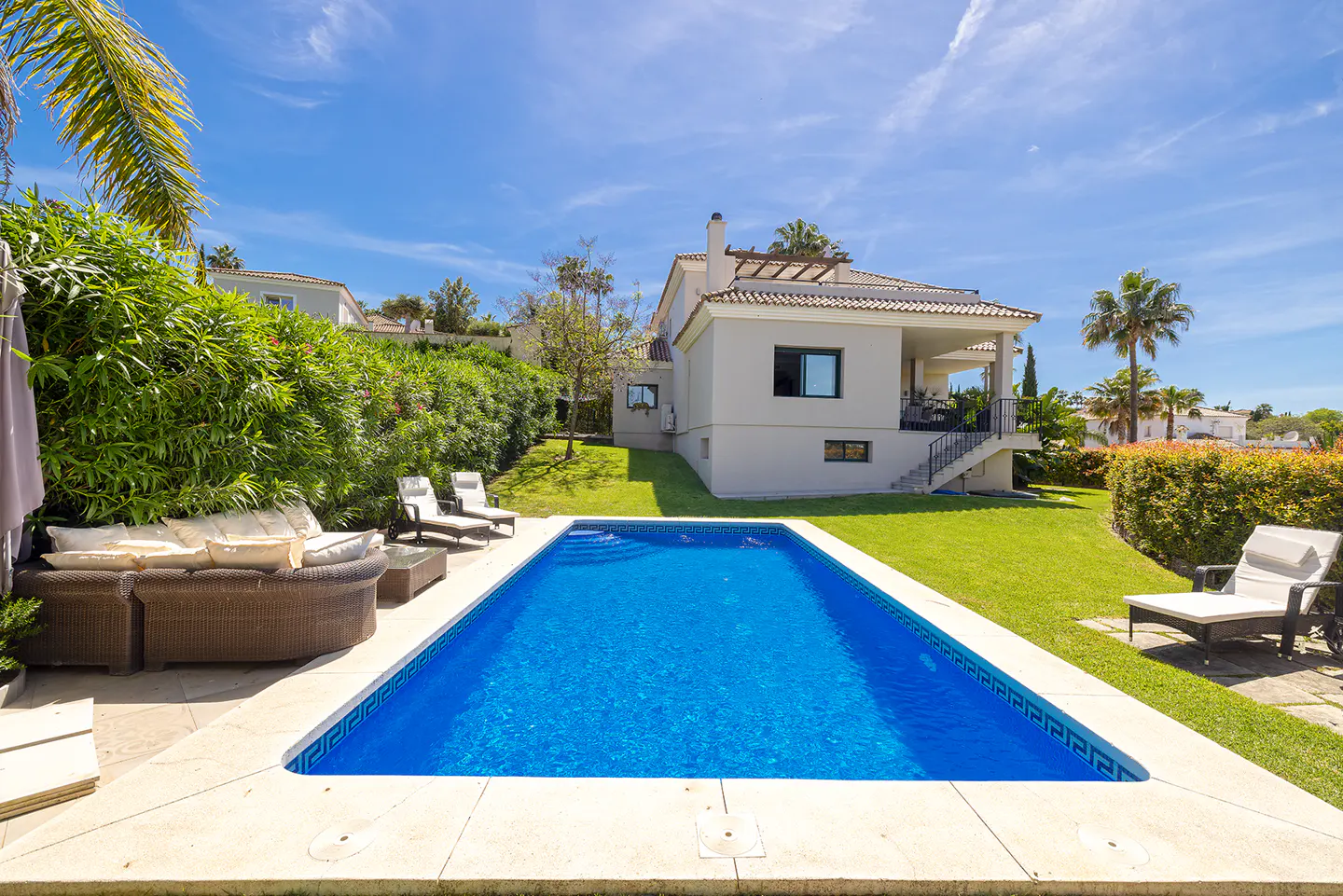 Backyard view of a beige house with a blue swimming pool, green grass, and outdoor furniture under a blue sky.