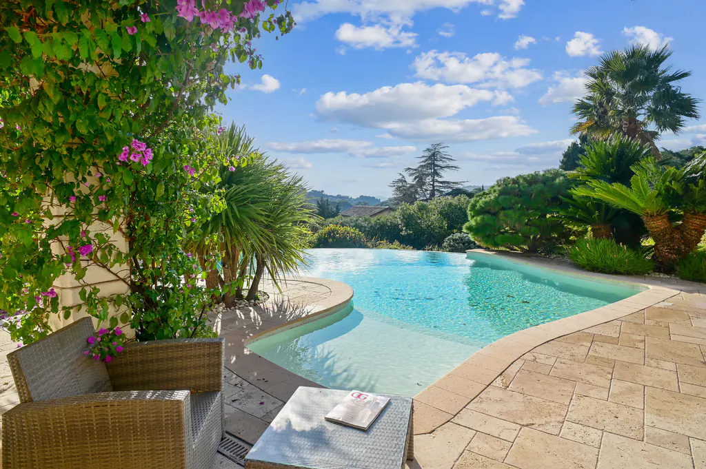 Outdoor patio with a pool, wicker furniture, and lush greenery under a blue sky with white clouds.