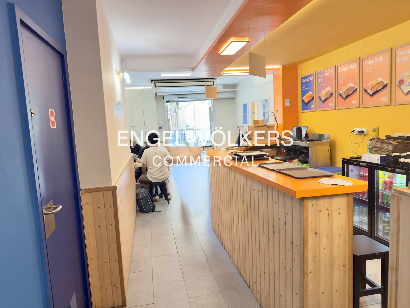 Restaurant interior with an orange counter, yellow walls with food posters, and people seated in the background.