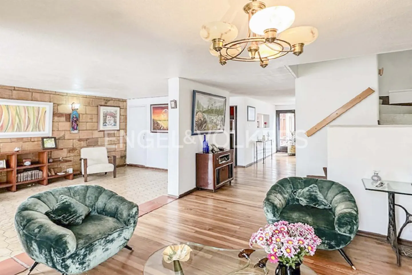 Bright living room with hardwood floors, green velvet chairs, and a glass coffee table with pink flowers. A chandelier hangs from the ceiling.