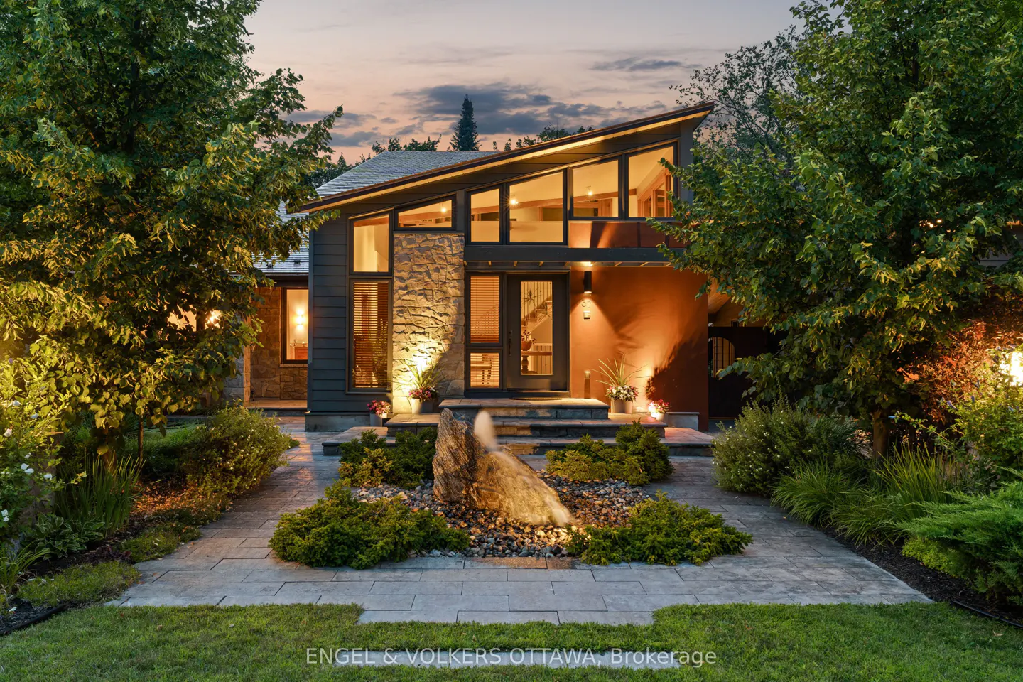 Exterior shot of a modern home with stone and dark siding, large windows, and a landscaped yard with a rock fountain.