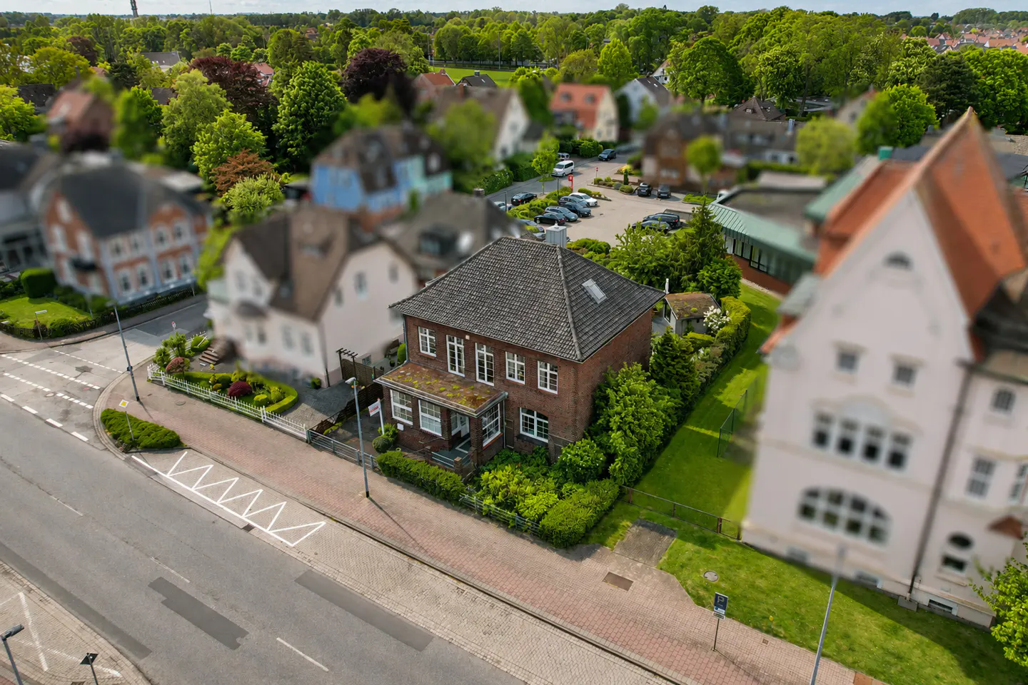 Aerial view of a two-story brick house with a gray roof, surrounded by green trees and lawns in a suburban neighborhood.