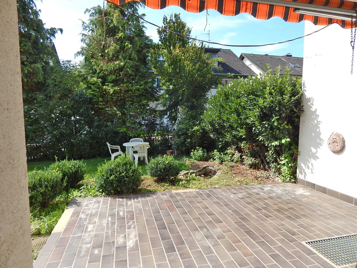 Patio with brick flooring leads to a green garden with a white table and chairs. An orange and white awning is overhead.