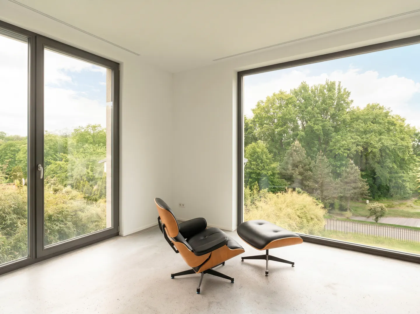 A modern lounge chair and ottoman in a bright, minimalist room with large windows overlooking a lush green forest.