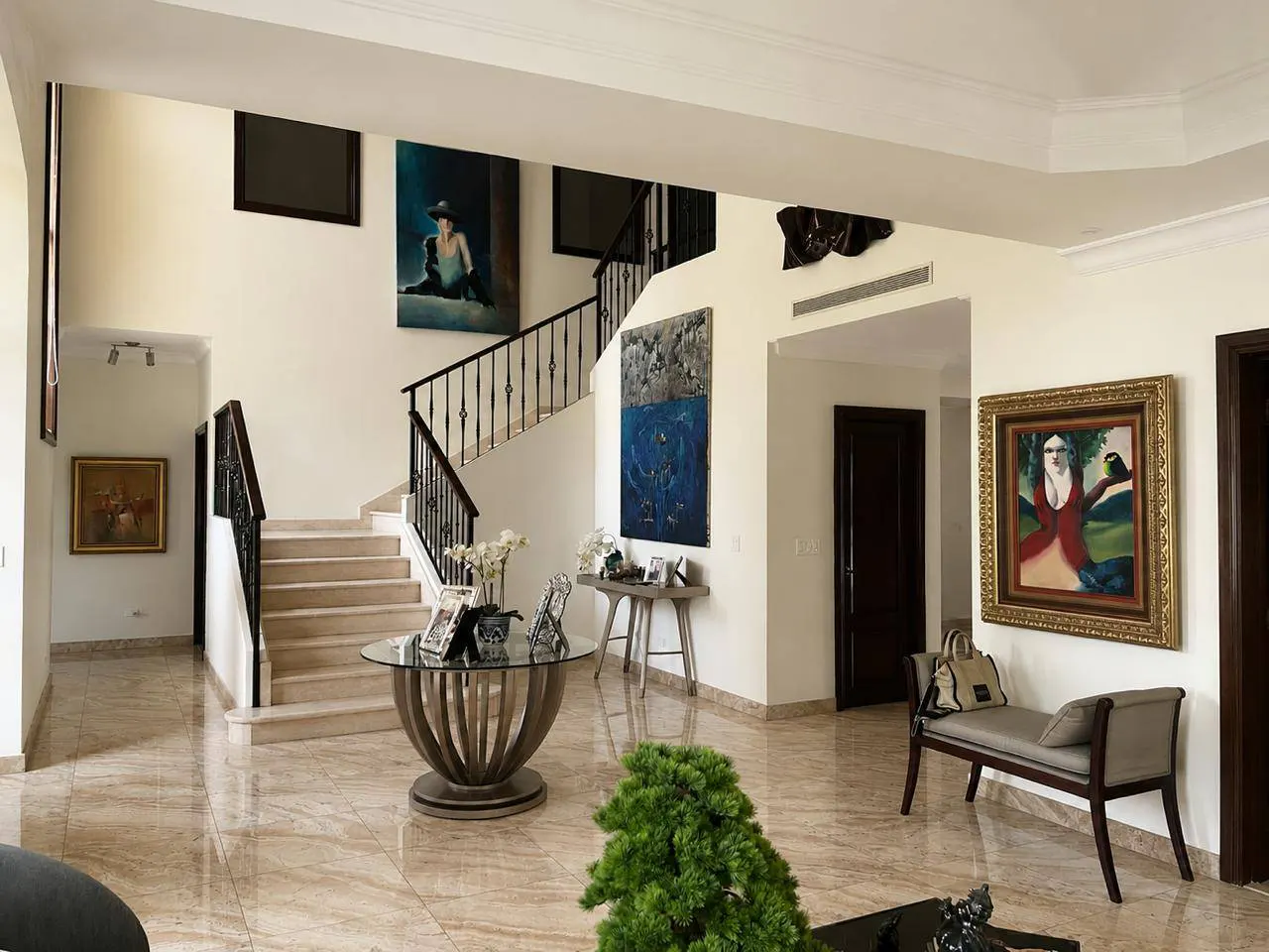 Bright foyer with marble floors, staircase, and artwork. A round glass table and bench seat add to the elegant decor.