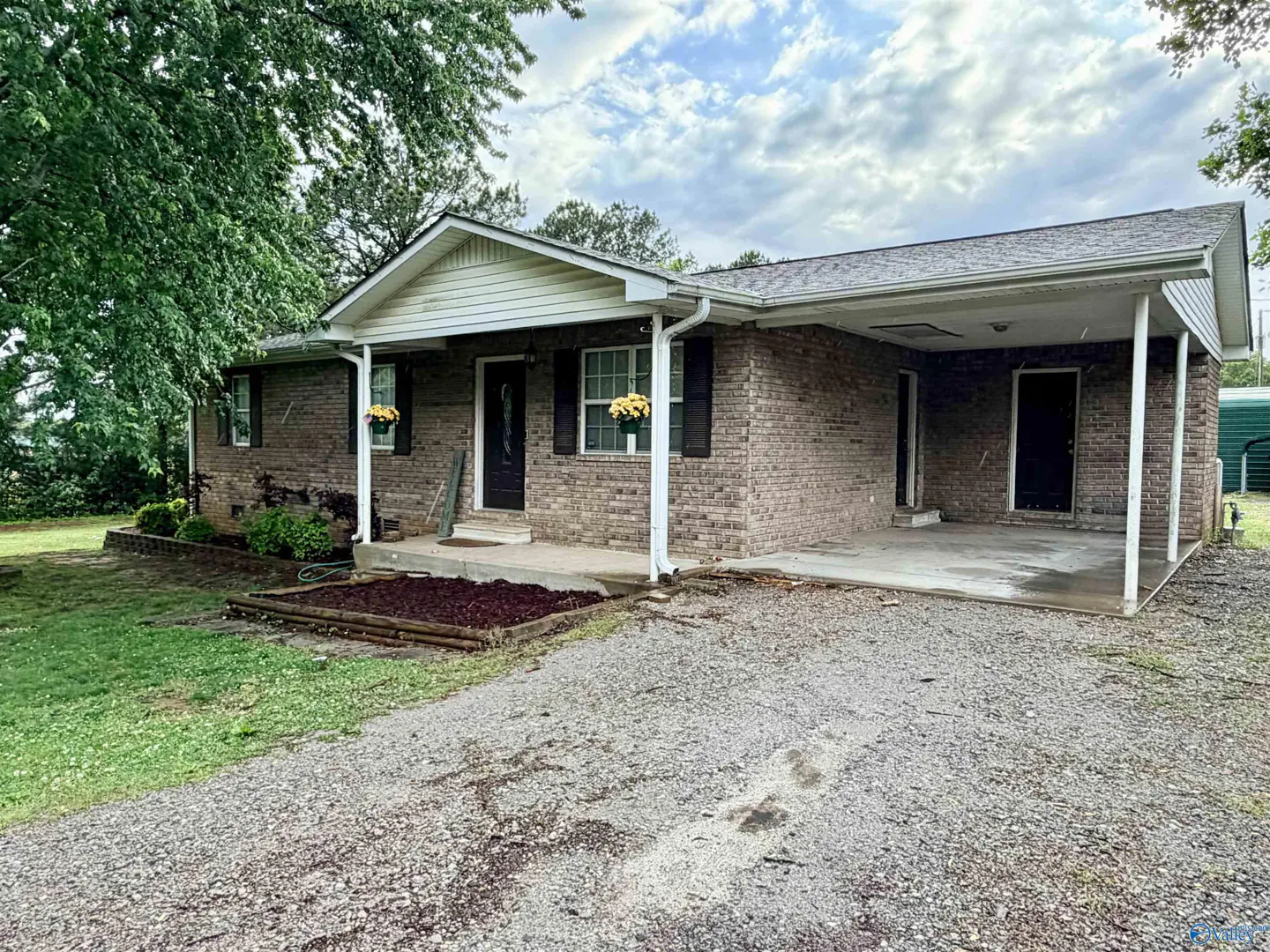 A single-story brick house with a carport, black shutters, and yellow flowers.