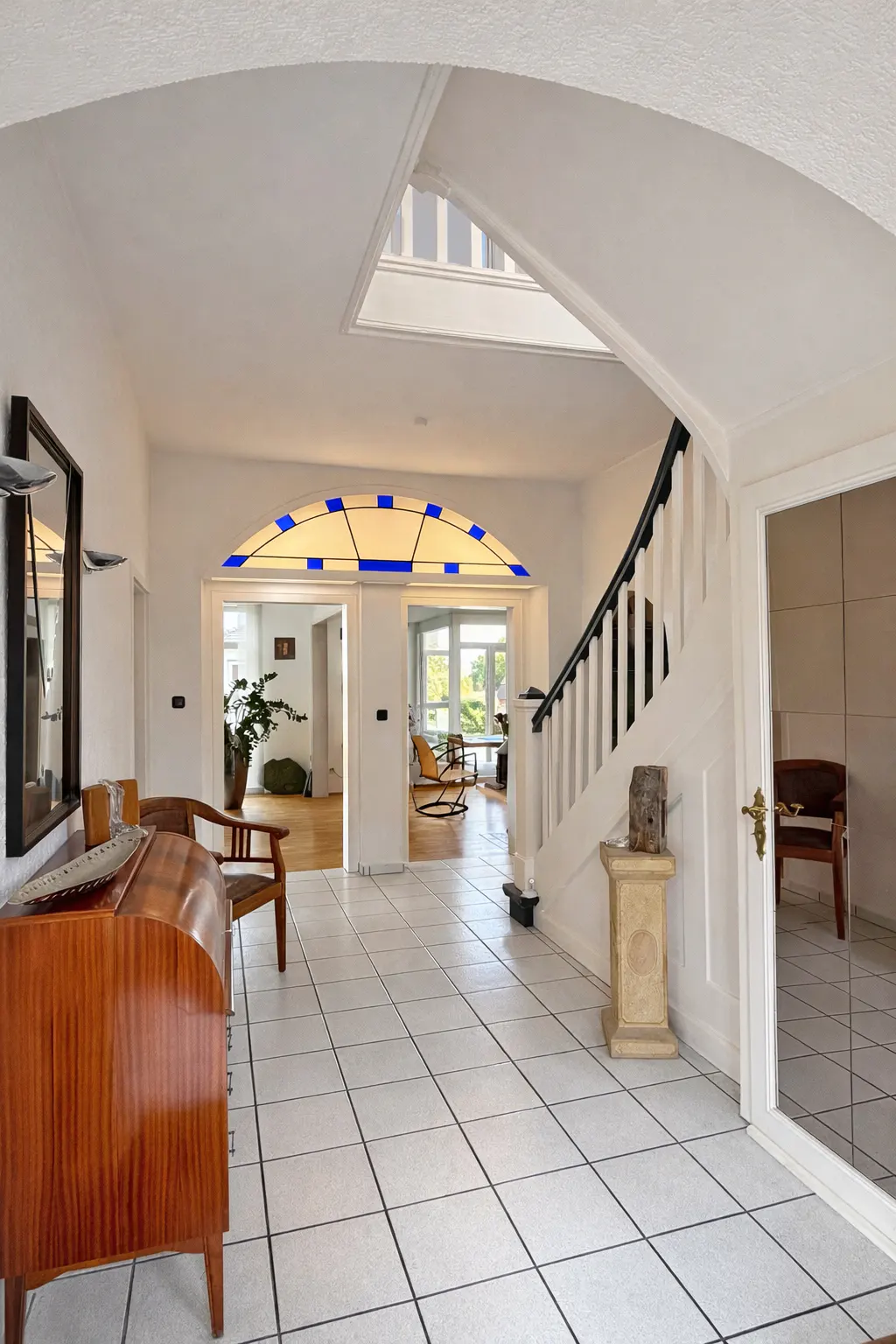 Bright foyer with white tile floor, arched doorways, and a staircase. A wooden dresser and chair are on the left.