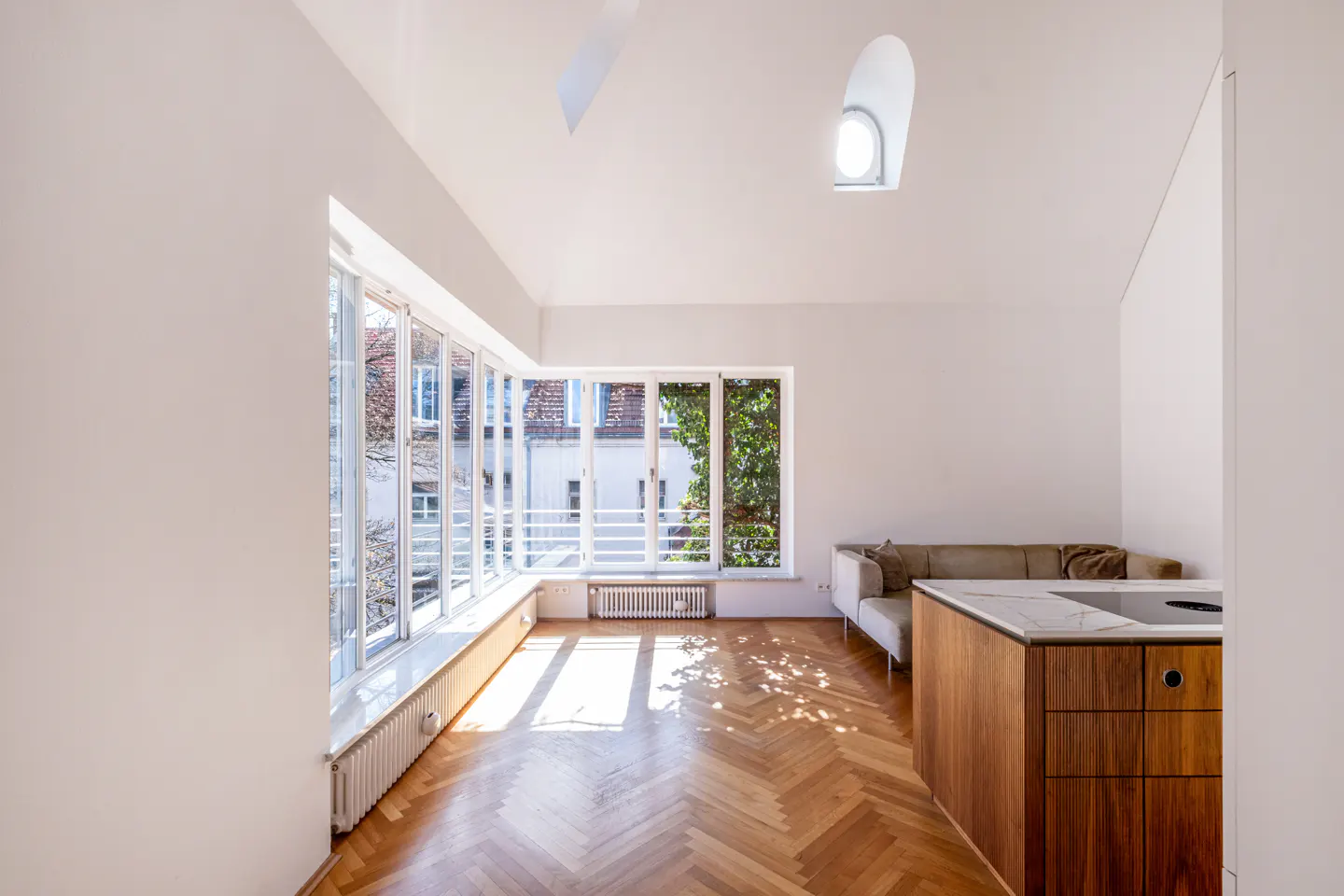 Bright, modern apartment interior with herringbone wood floors, white walls, and large windows. A kitchen island and sofa are visible.