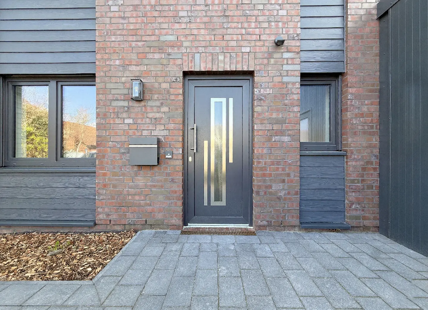 Modern home exterior with a gray front door, brick facade, and gray wood siding. A gray mailbox is mounted next to the door.