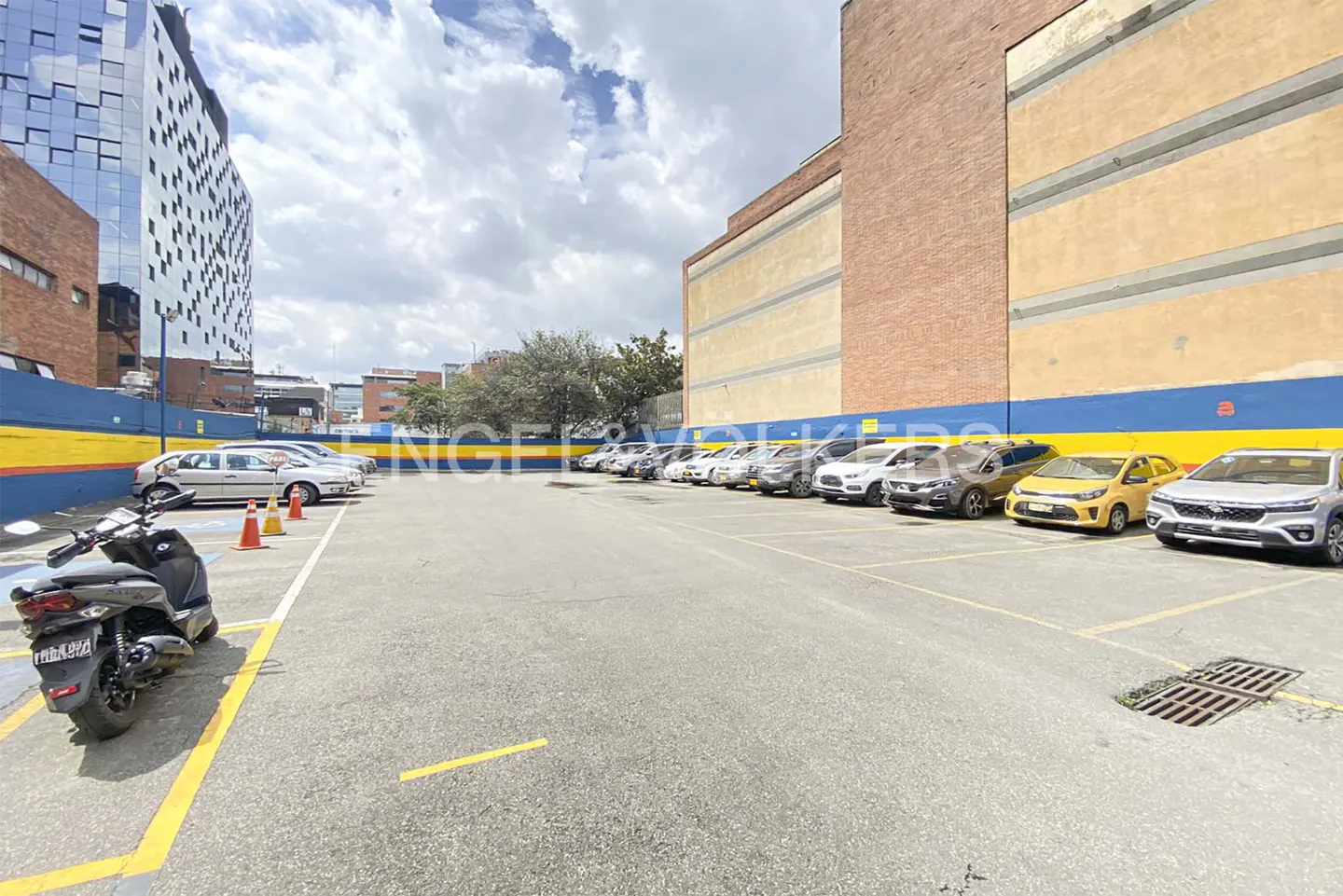 Outdoor parking lot with rows of parked cars, a scooter in the foreground, and a large brick building under a cloudy sky.
