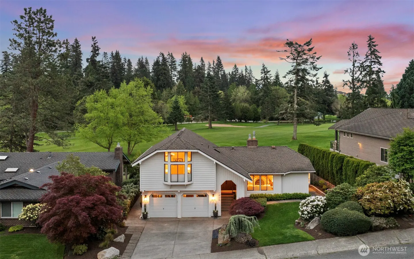 Two-story white house with a two-car garage, green lawn, and trees in the background under a colorful sunset sky.