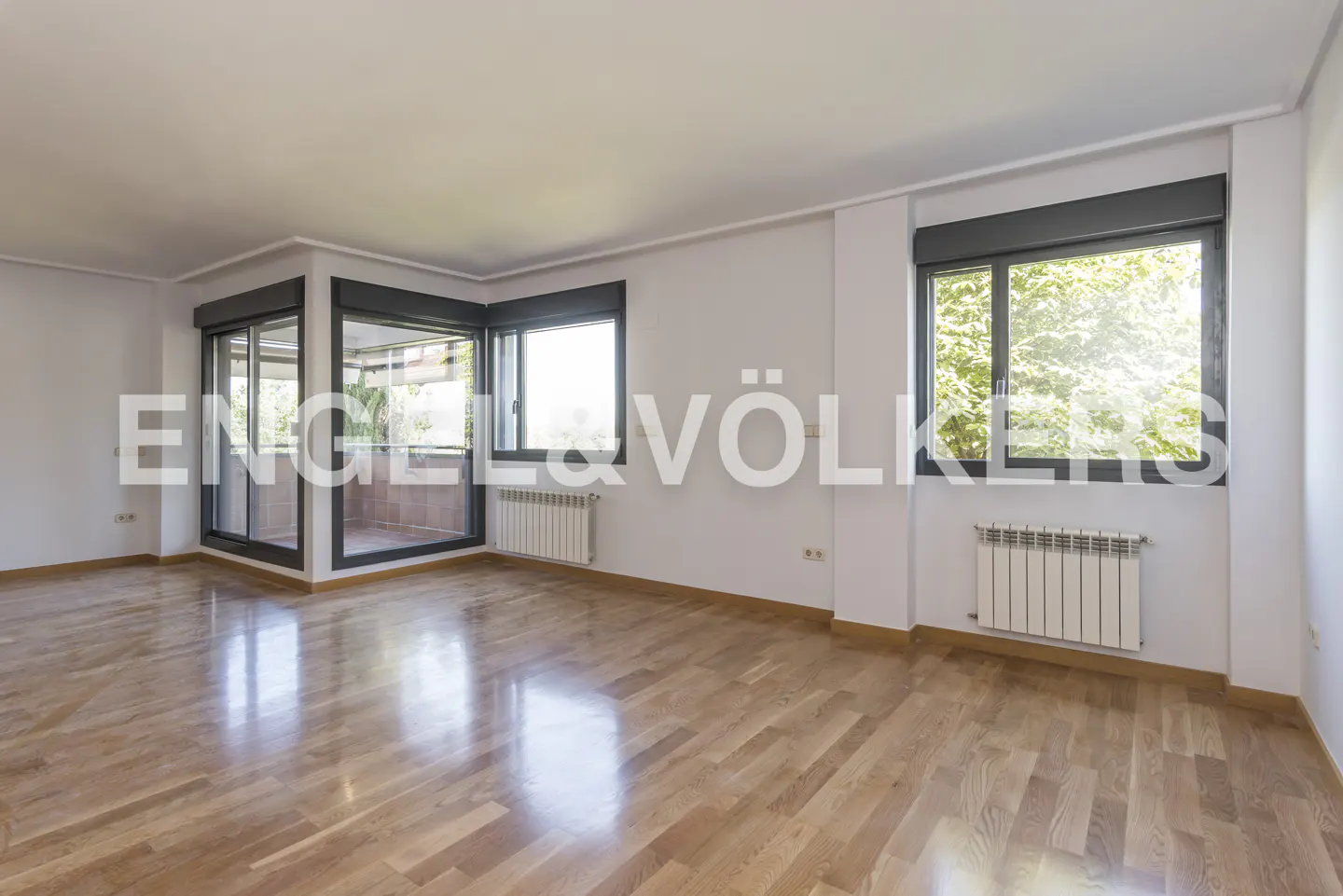 Bright, empty room with wood floors, white walls, and large windows with dark frames. Radiators are under the windows. Balcony visible through glass doors.