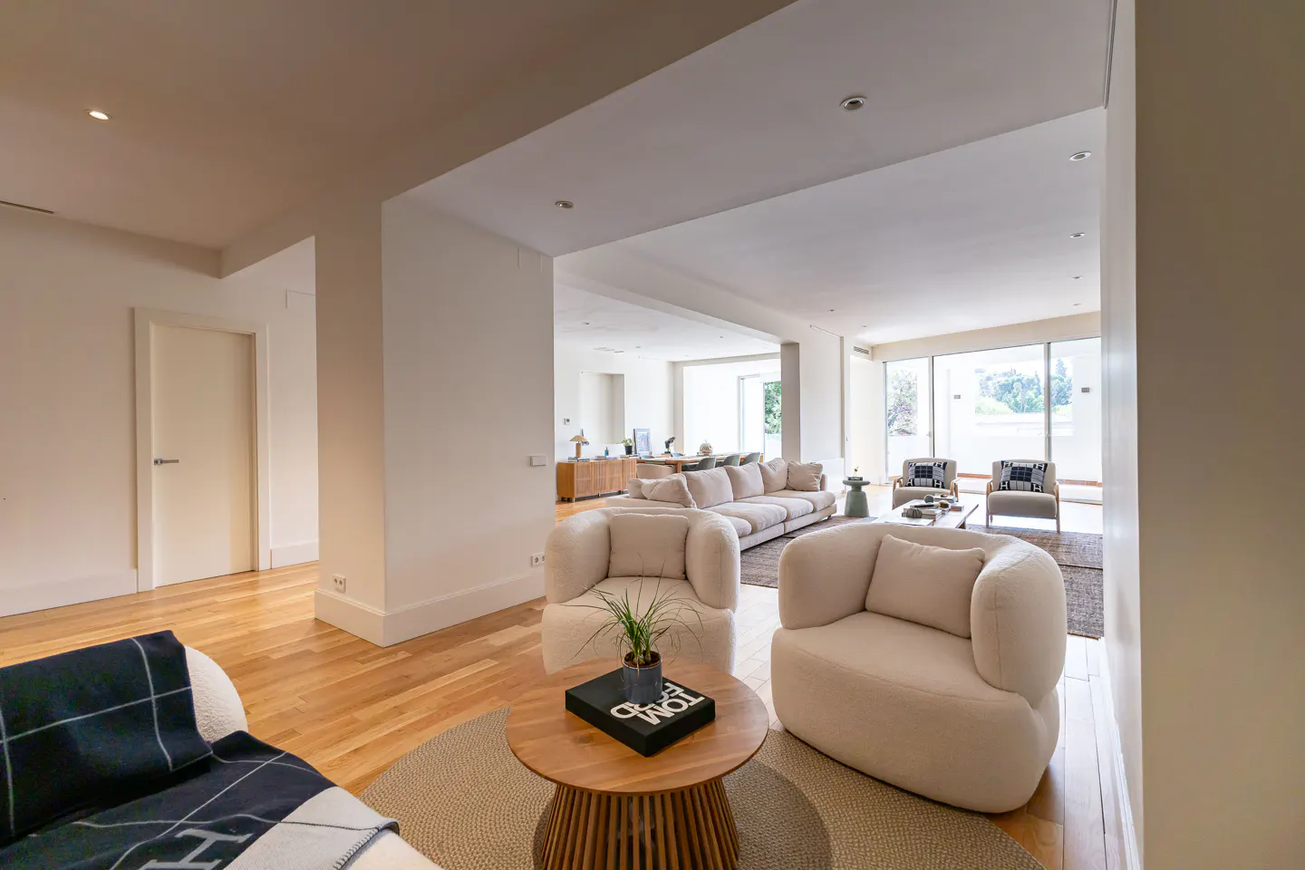 Bright, open-concept living room with hardwood floors. Two cream-colored armchairs flank a round wood table with a plant. A large sofa is visible in the background.