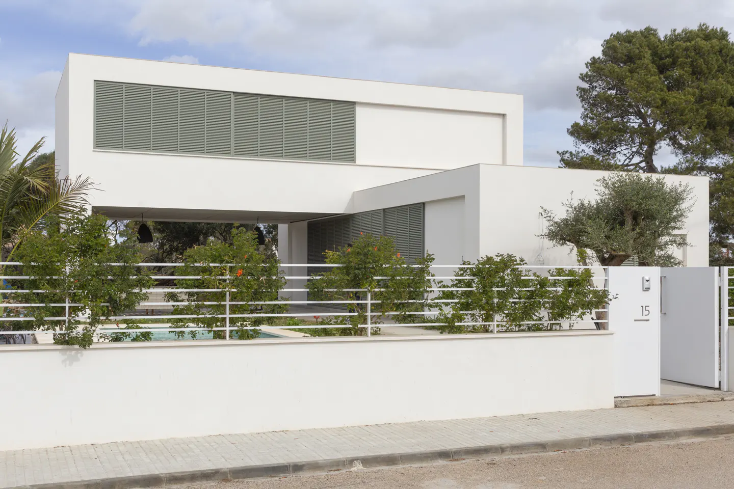 Modern white two-story house with horizontal window blinds, white fence, and green bushes.