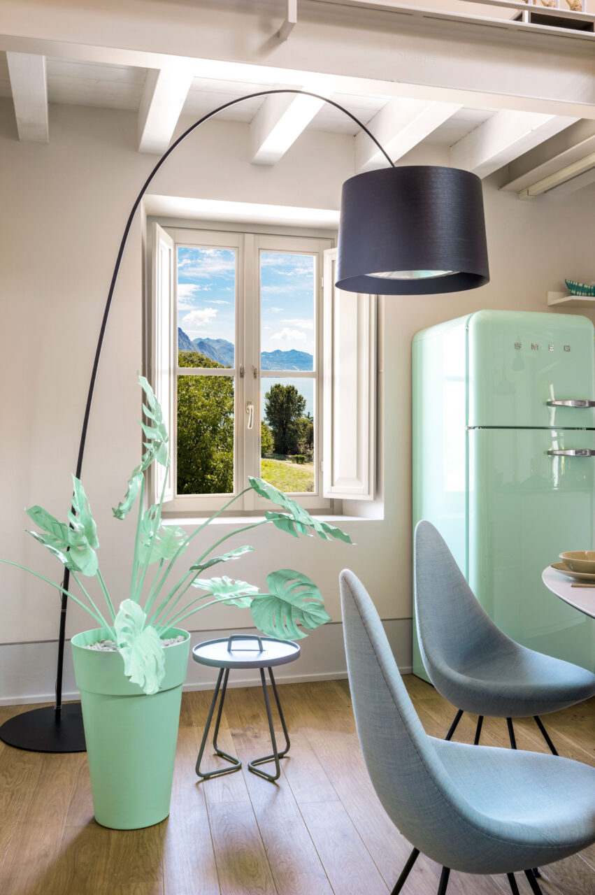 Interior view of a room with a mint green color scheme, featuring a large plant, chairs, and a retro-style refrigerator. A window overlooks a scenic landscape.
