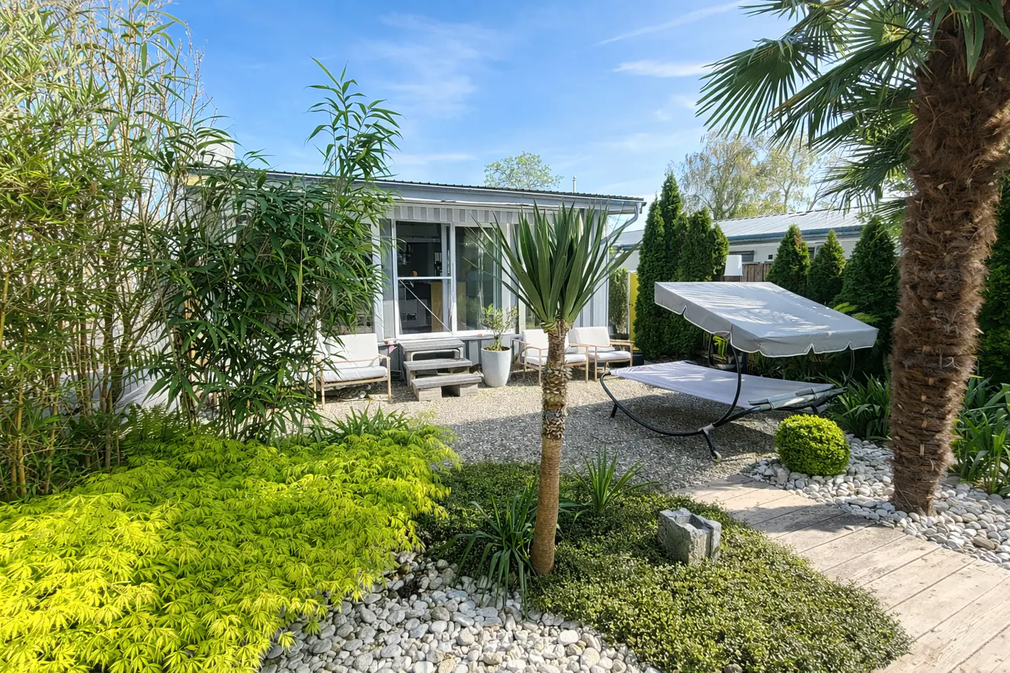 A sunny patio with a gravel floor, modern outdoor chairs, a canopy daybed, and lush tropical landscaping.