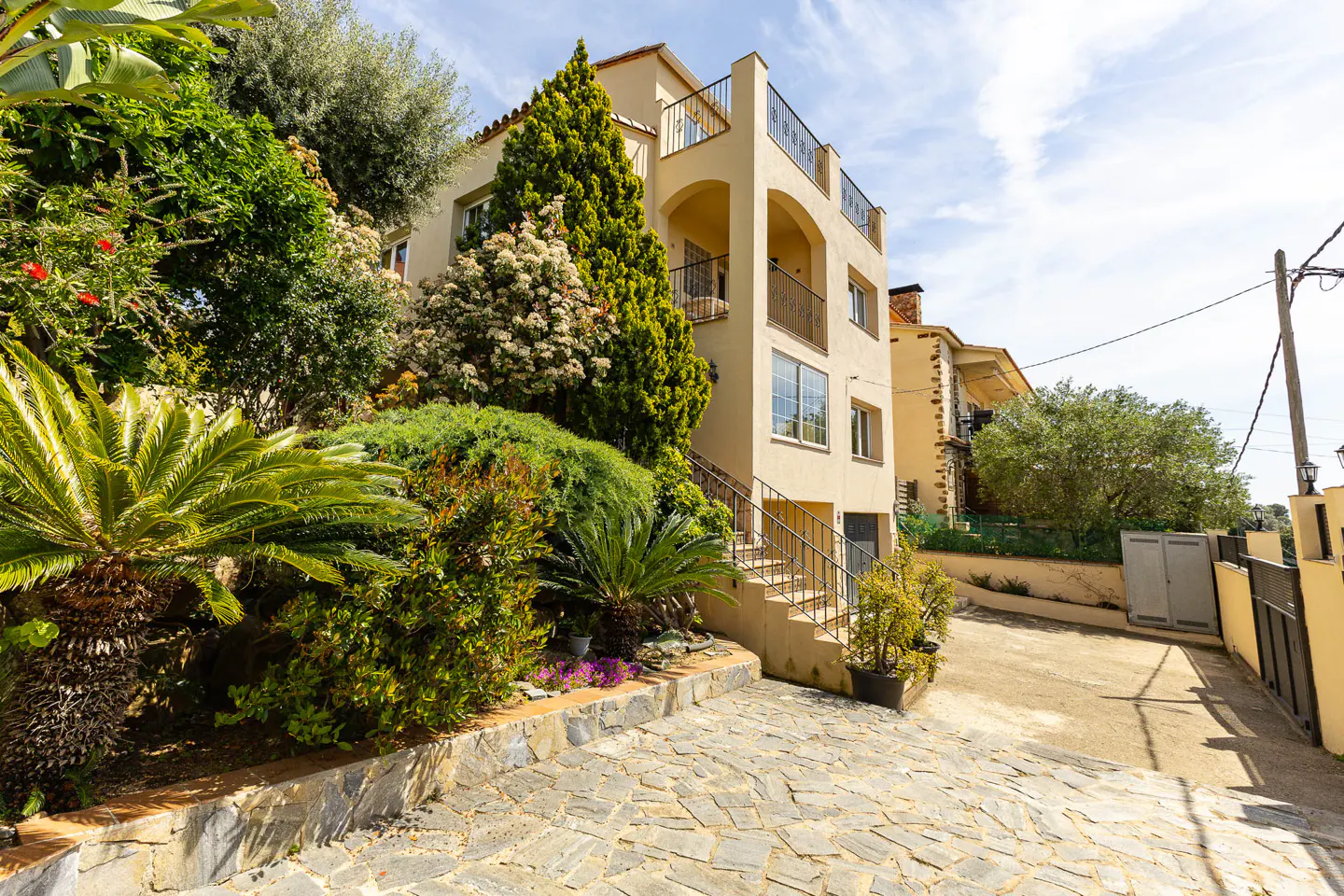 A tan, multi-story house with balconies and lush greenery. A stone driveway leads up to the house.
