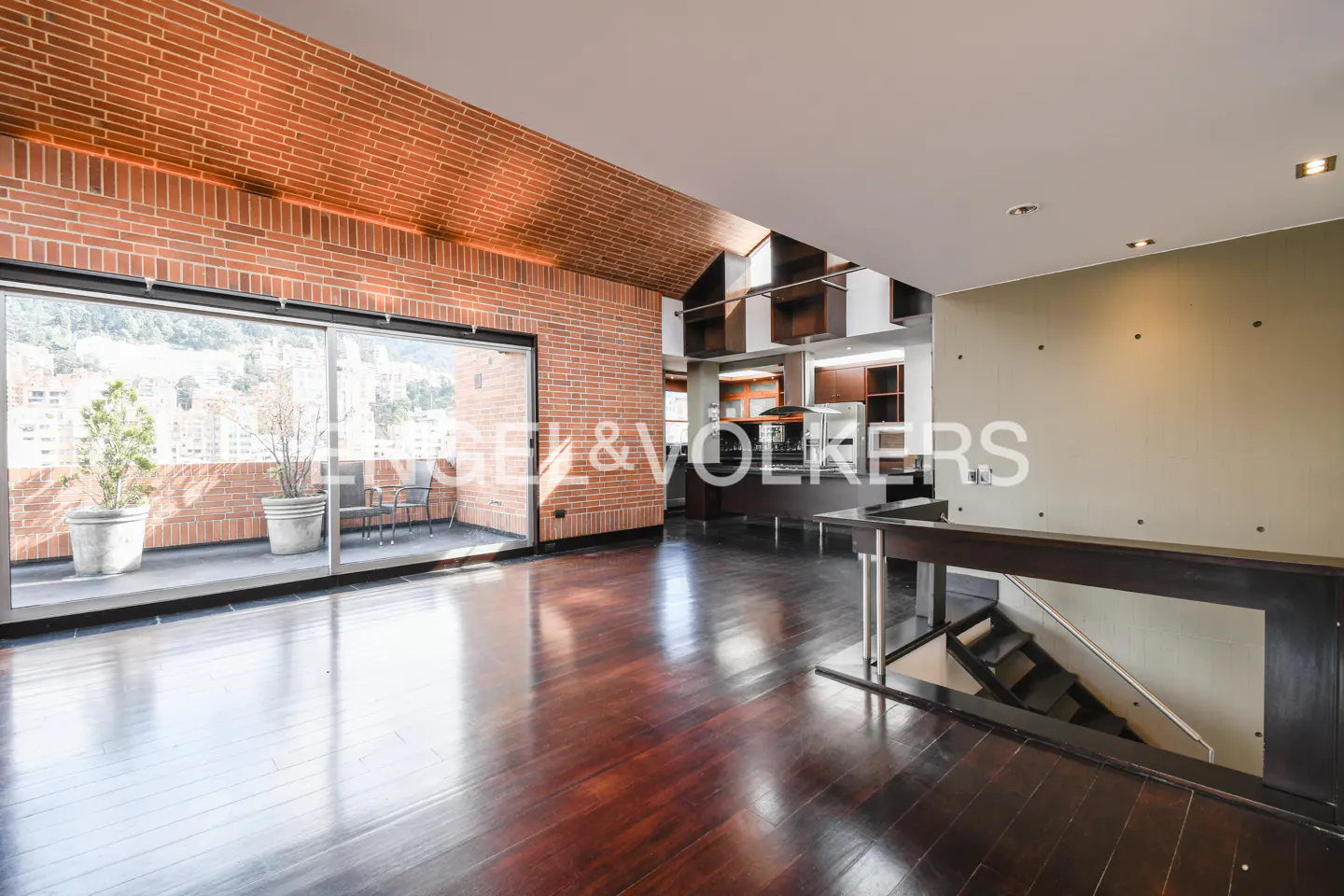 Open-concept living space with brick walls, dark wood floors, and a balcony with city views. Stairs lead to another level.