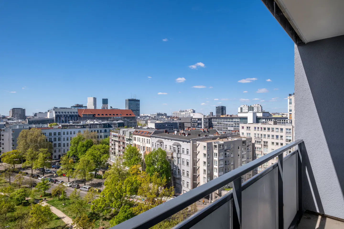 View from a balcony with gray railings overlooking a city with green trees under a blue sky.