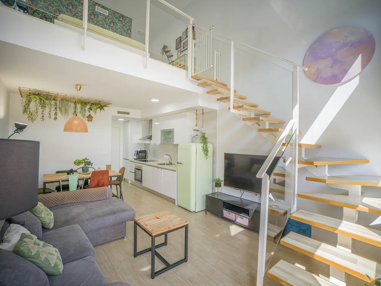 Bright, modern apartment interior with a gray sofa, dining table, kitchen, TV, and wooden stairs leading to a loft. A light green fridge adds a pop of color.