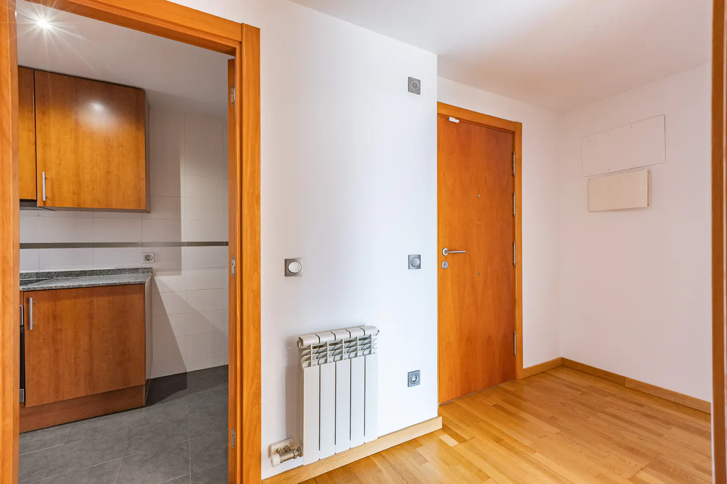 A hallway with light wood floors, a white wall with a radiator, and a doorway leading to a kitchen with wood cabinets.