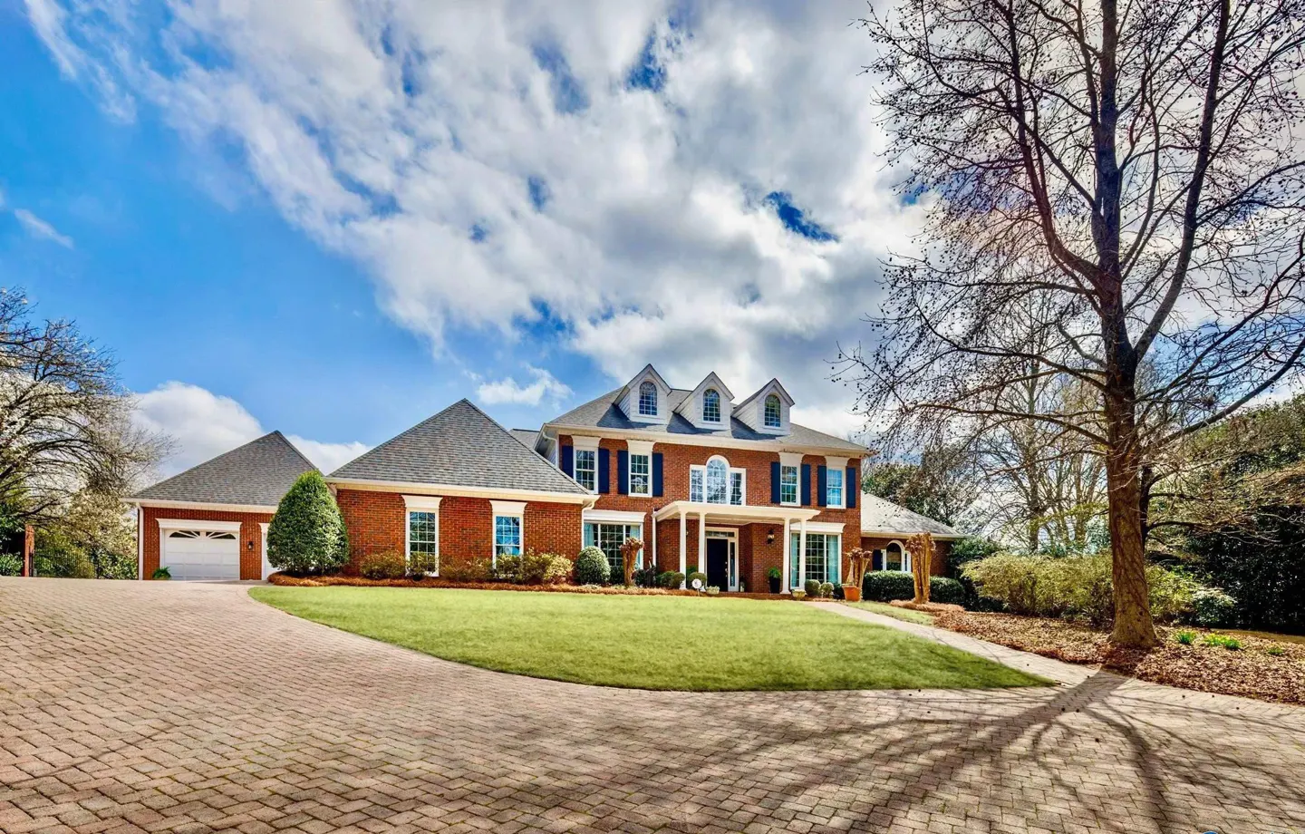 Brick house with blue shutters, white trim, and a gray roof, set on a green lawn under a blue sky with clouds.