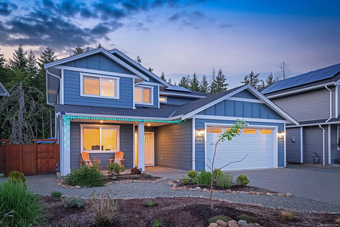 Two-story blue house with white trim, a white garage door, and a gravel yard with plants. The sky is blue with clouds.