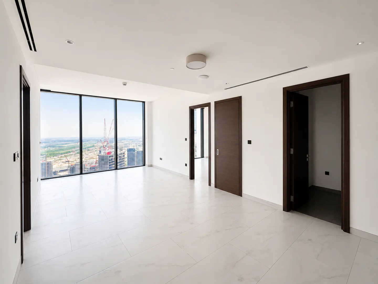 Bright, empty room with white walls and marble floors. Large window shows a city skyline with construction. Three dark wood doors are visible.