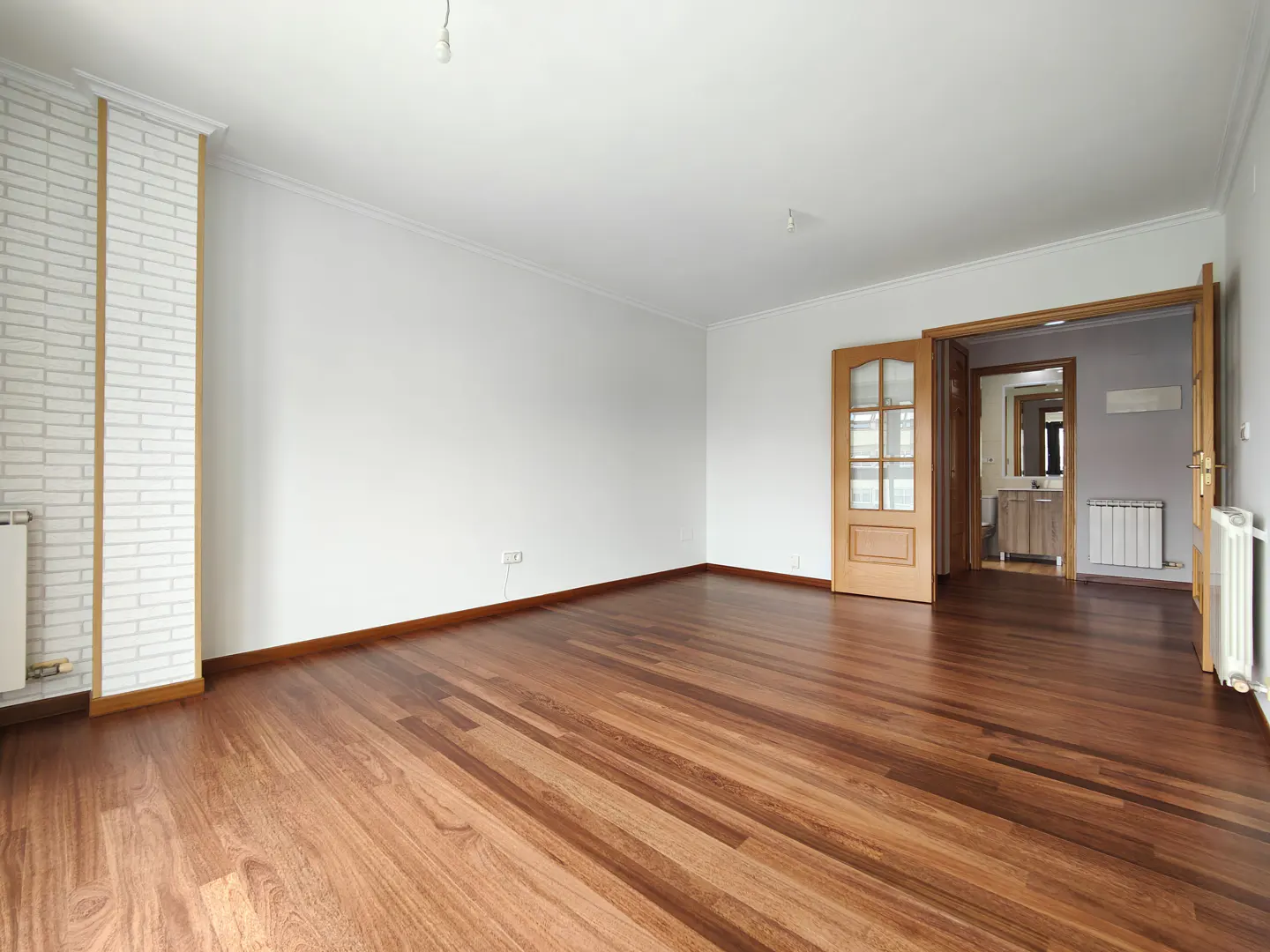 Empty room with white walls, a white brick accent pillar, and polished dark wood flooring, leading into a bathroom.